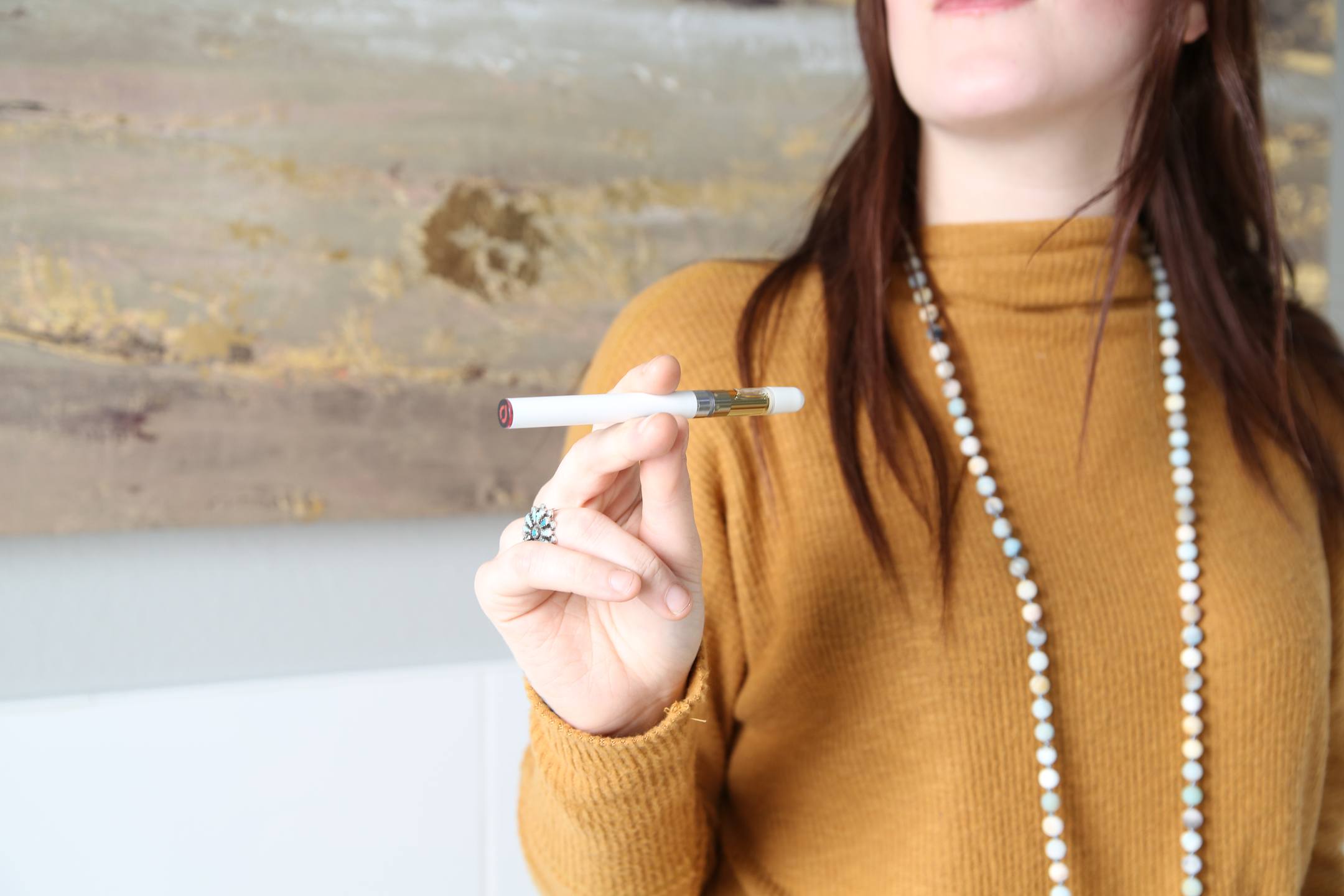 A brunette woman in an orange sweater and long pearl necklace holds an elegant Joy Organics CBD vape pen in one hand.