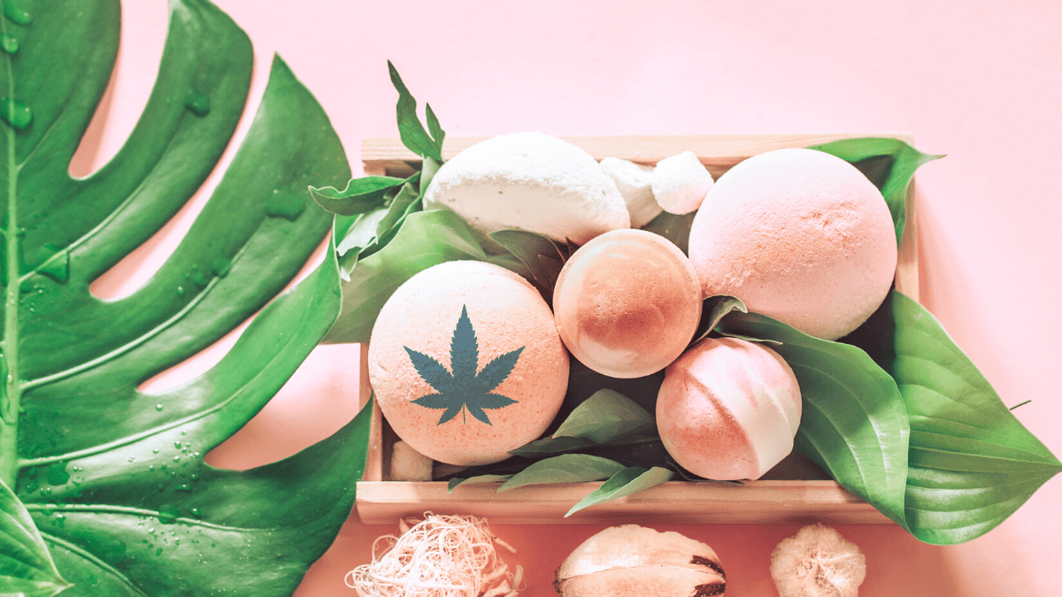 We reviewed the best CBD bath bombs and soaks. A photo of pink bath bombs artfully arranged in a box with sea shells and big green leaves. One bath bomb is digitally imprinted with a hemp leaf.