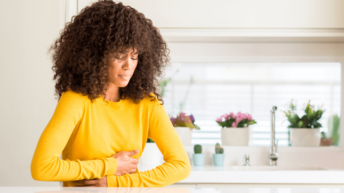 Can CBD help ease nausea? Photo: A black woman with natural hair, wearing a yellow sweater and standing in a kitchen, holds her stomach as if nauseous or in stomach pain.